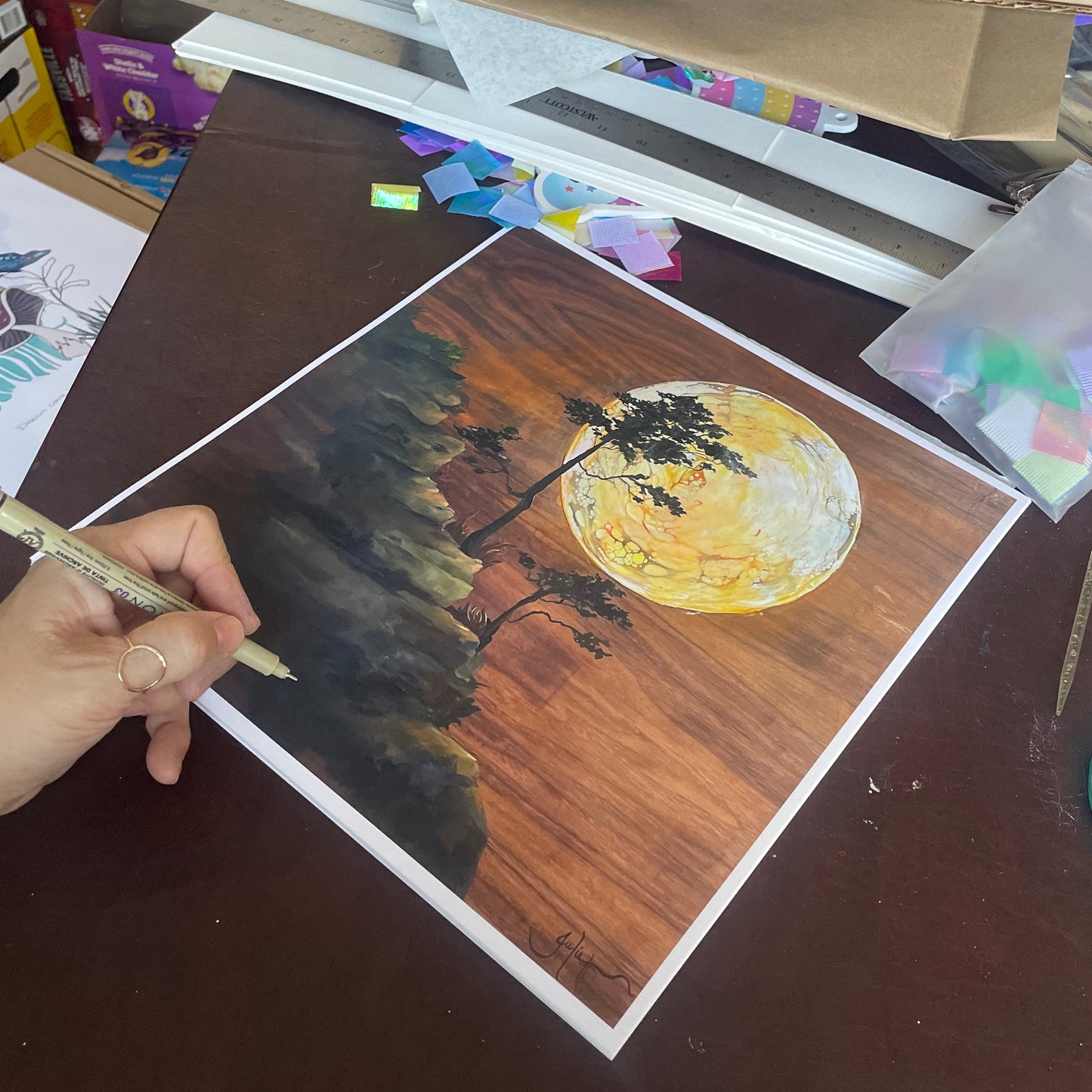 Artistic depiction of a landscape with a tree and moon on a table with art supplies.